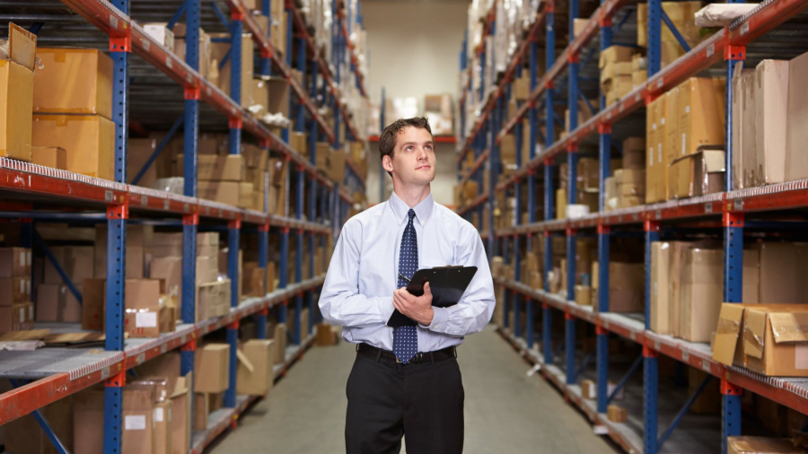 A man in business attire holds a clipboard while standing in the aisle of a warehouse, surrounded by tall shelves filled with cardboard boxes.