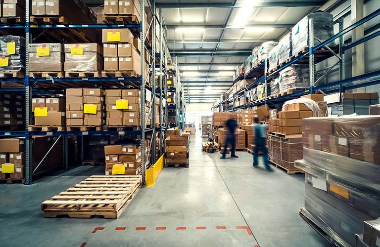 Large warehouse interior with tall shelves stacked with cardboard boxes. Two workers move through the aisle, with pallets and wrapped goods visible on the floor. The area is well-lit with overhead lights.