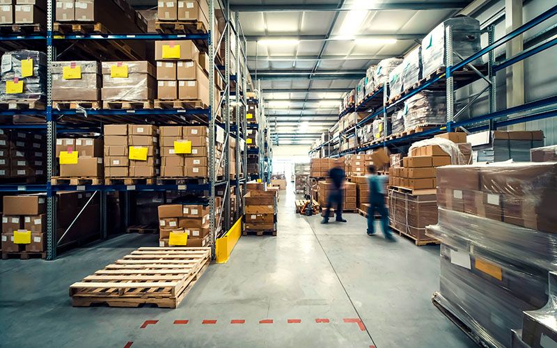 Large warehouse interior with tall shelves stacked with cardboard boxes. Two workers move through the aisle, with pallets and wrapped goods visible on the floor. The area is well-lit with overhead lights.