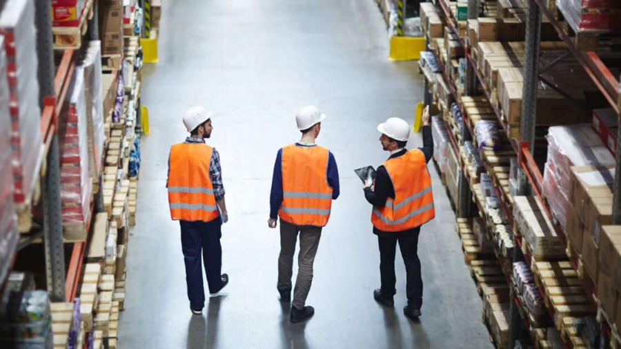 Three workers wearing orange safety vests and white hard hats walk down an aisle in a warehouse, surrounded by shelves stocked with various boxes and products. One person is holding a clipboard.