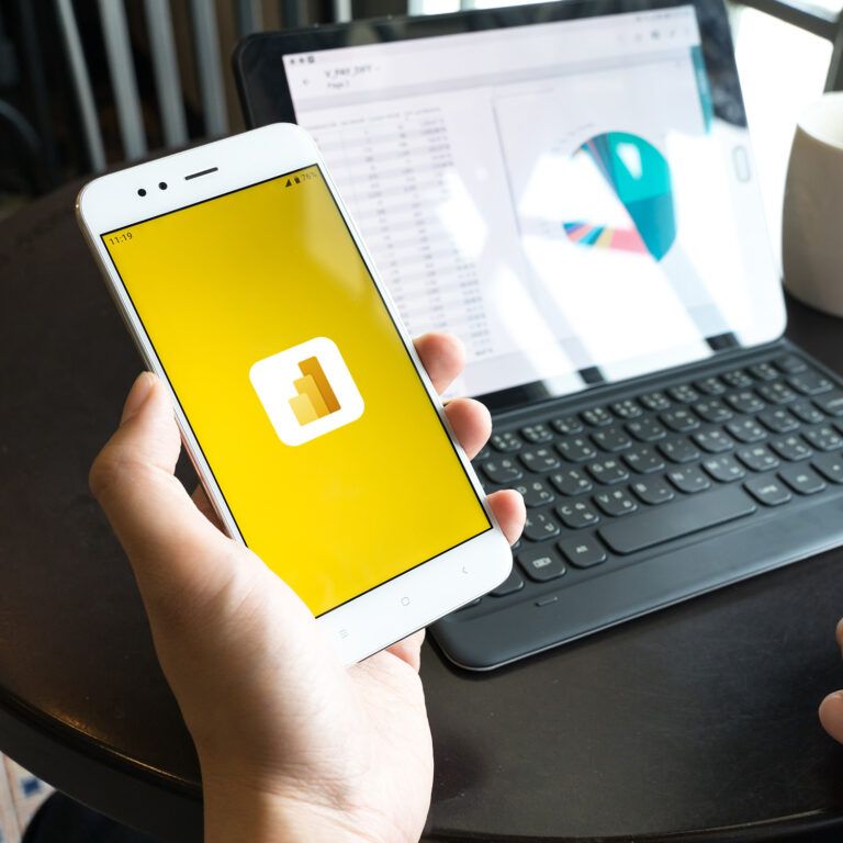 A person holds a smartphone displaying the Power BI logo, while a tablet with a keyboard on a round table shows data charts; there is also a white coffee mug beside the tablet.
