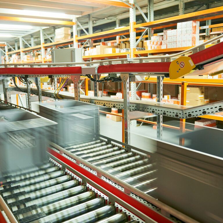 Plastic bins move quickly along a conveyor belt system in a warehouse, with shelves of boxes and packages in the background—showcasing an automated logistics center ideal for advanced automation analysis.