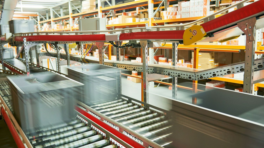 Plastic bins move quickly along a conveyor belt system in a warehouse, with shelves of boxes and packages in the background—showcasing an automated logistics center ideal for advanced automation analysis.