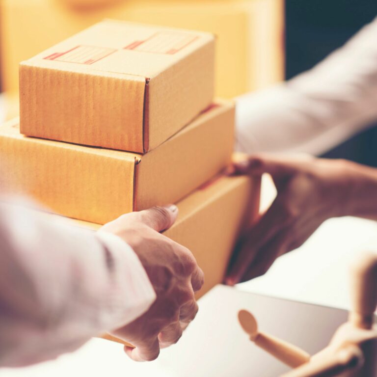 Two people are exchanging a stack of three small cardboard boxes. One person is handing the boxes over while the other is receiving them. The background is blurred.