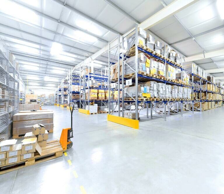 Spacious warehouse with tall metal shelves stocked with boxes and pallets; an orange pallet jack and stacked goods are visible in the foreground. The space is brightly lit and organized.