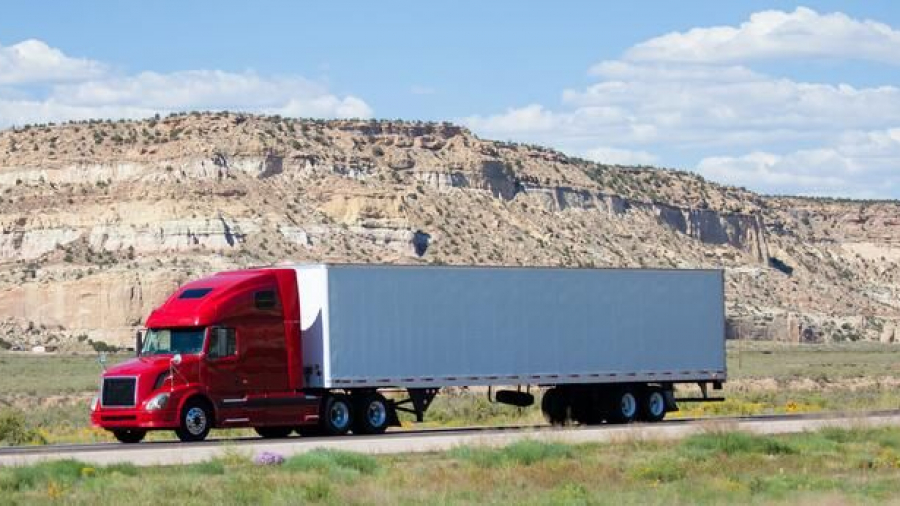 A bright red semi-truck with a long, gray trailer drives along a highway in a desert landscape, with rocky hills and a partly cloudy sky in the background.