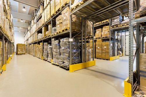 Warehouse interior with tall shelves stacked with cardboard boxes and packages. The floor is clear and clean, with wide aisles for movement. The area is well-lit with overhead lights.