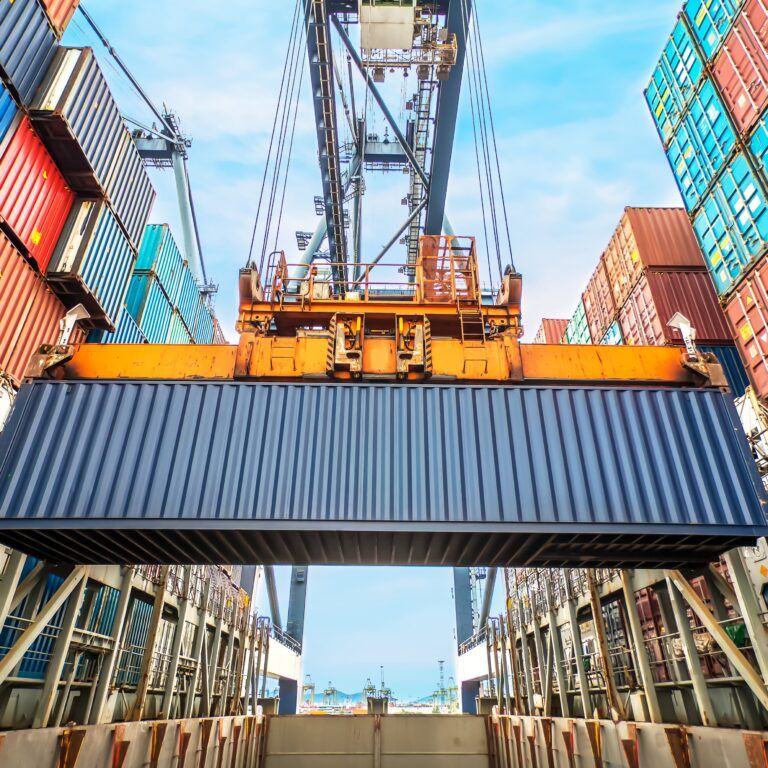 A large crane lifts a blue shipping container in a busy port, surrounded by stacks of colorful containers under a blue sky.