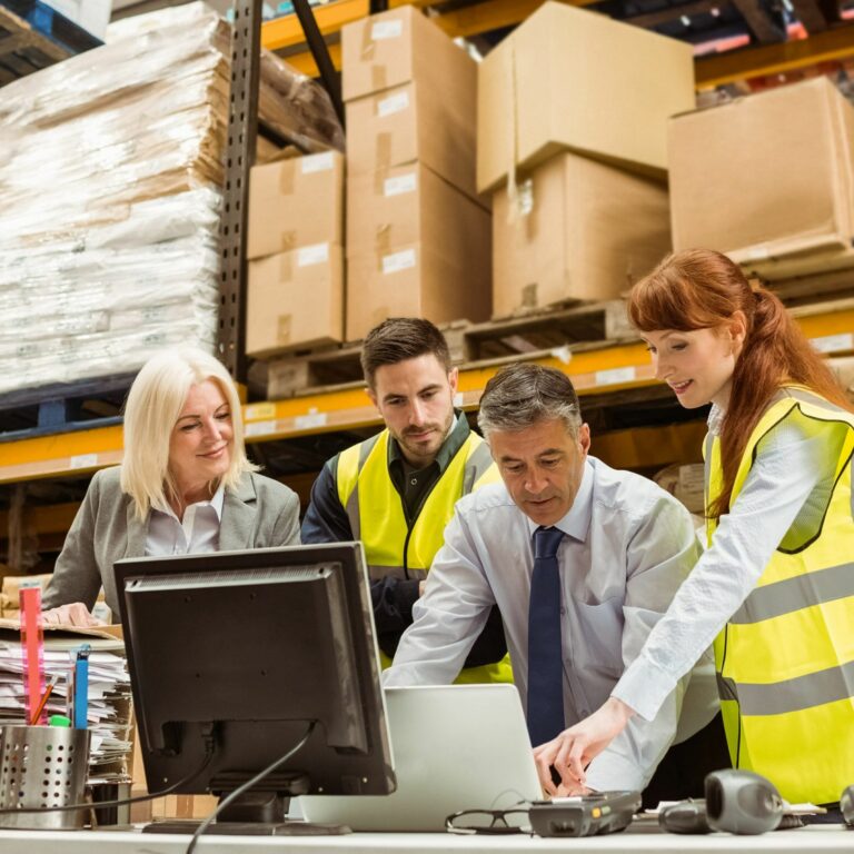 Four people in a warehouse, wearing high-visibility vests, gather around a desk with a computer and paperwork, collaborating on a project with shelves of boxes and packages in the background.