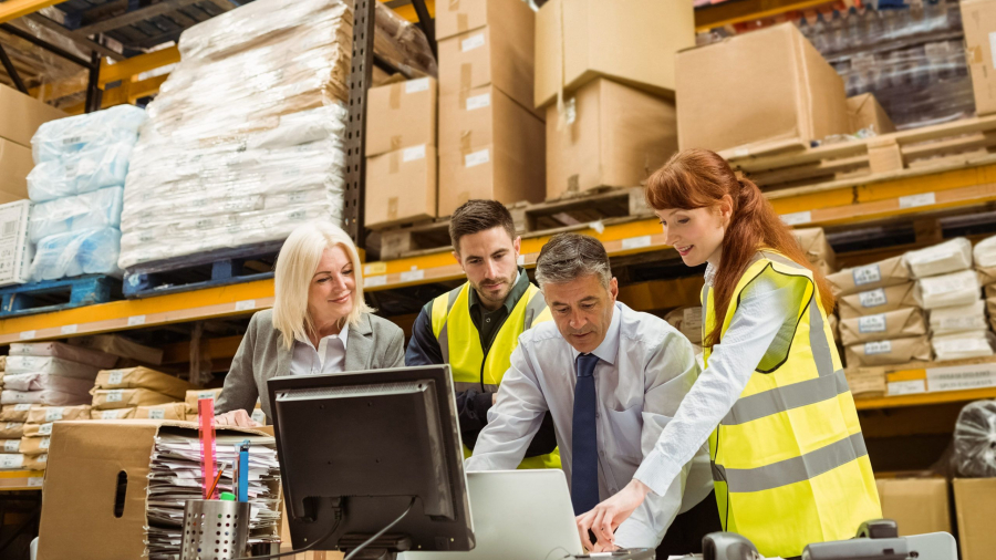 Four people in a warehouse, wearing high-visibility vests, gather around a desk with a computer and paperwork, collaborating on a project with shelves of boxes and packages in the background.