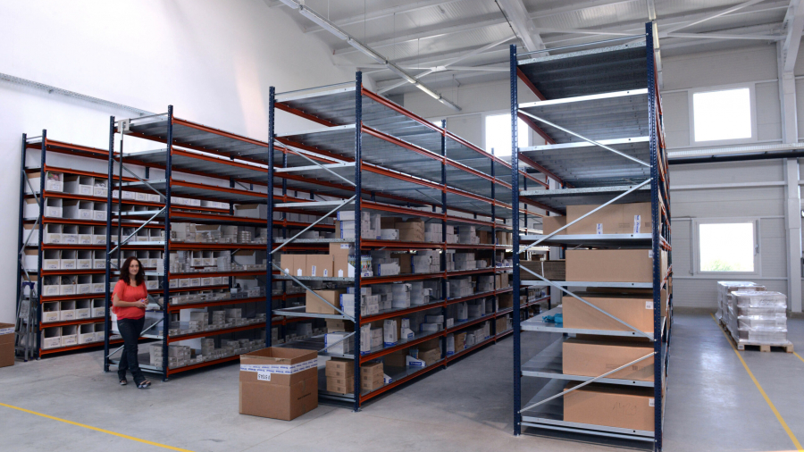 A woman walks through a spacious warehouse with tall metal shelves holding various boxes and items. The area is well-lit with high ceilings and large windows. Some boxes are stacked on the floor.