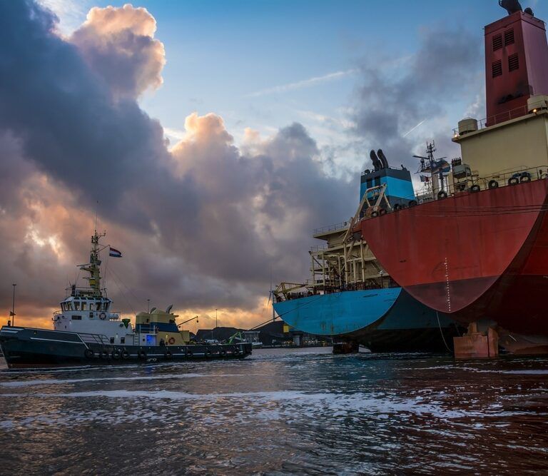 A tugboat guides a large cargo ship near a dock at sunset, with dramatic clouds in the sky and reflections on the water. The cargo ship’s hulls are red and blue, showing signs of wear.