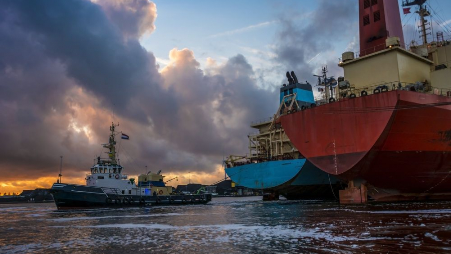 A tugboat guides a large cargo ship near a dock at sunset, with dramatic clouds in the sky and reflections on the water. The cargo ship’s hulls are red and blue, showing signs of wear.