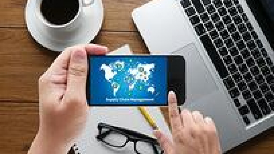 A person holds a smartphone displaying a supply chain management graphic, with a laptop, coffee cup, notebook, and glasses on a wooden desk in the background.