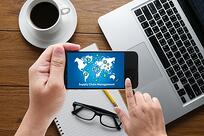 A person holds a smartphone displaying a supply chain management graphic, with a laptop, coffee cup, notebook, and glasses on a wooden desk in the background.