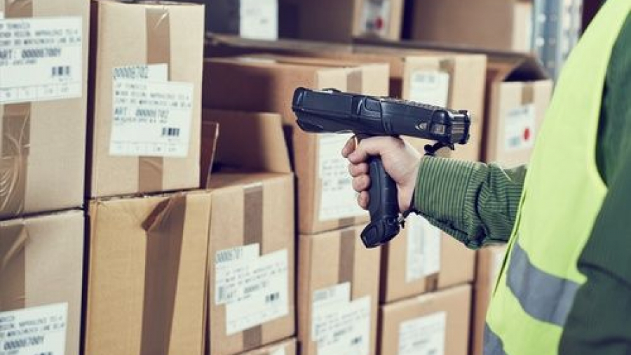 A person in a high-visibility vest scans barcodes on stacked cardboard boxes in a warehouse using a handheld barcode scanner.