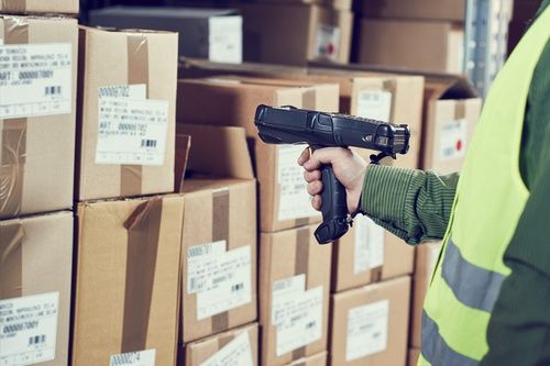A person in a high-visibility vest scans barcodes on stacked cardboard boxes in a warehouse using a handheld barcode scanner.
