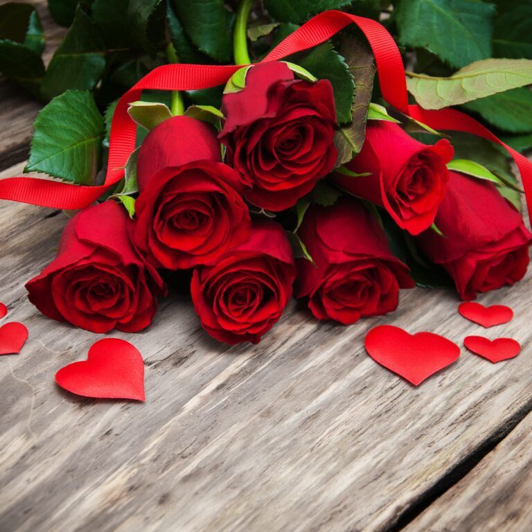A bouquet of red roses with green leaves is placed on a wooden surface. Red paper hearts and a red ribbon are scattered around the flowers, creating a romantic atmosphere.