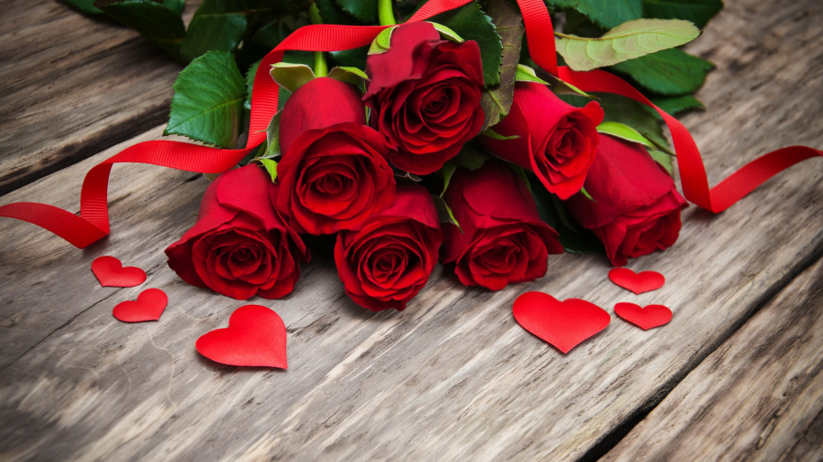 A bouquet of red roses with green leaves is placed on a wooden surface. Red paper hearts and a red ribbon are scattered around the flowers, creating a romantic atmosphere.