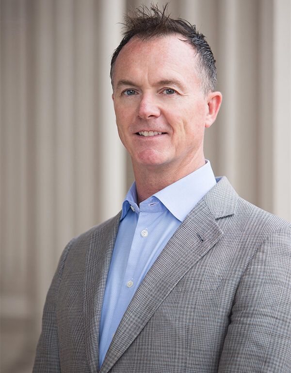 A man wearing a light gray suit jacket and a light blue shirt smiles with confidence, embodying leadership as he stands in front of a background with tall, blurred columns.