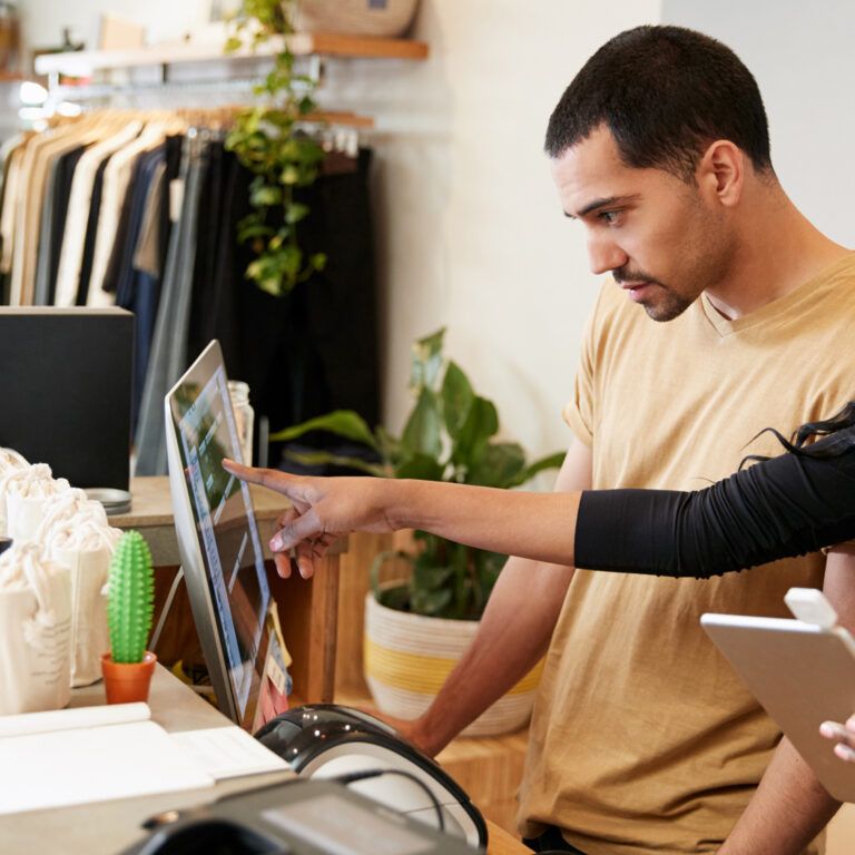 Two people stand behind a clothing store counter, looking at a computer screen. One points at the monitor while holding a tablet; the other watches attentively, demonstrating how retail management systems streamline their workflow.