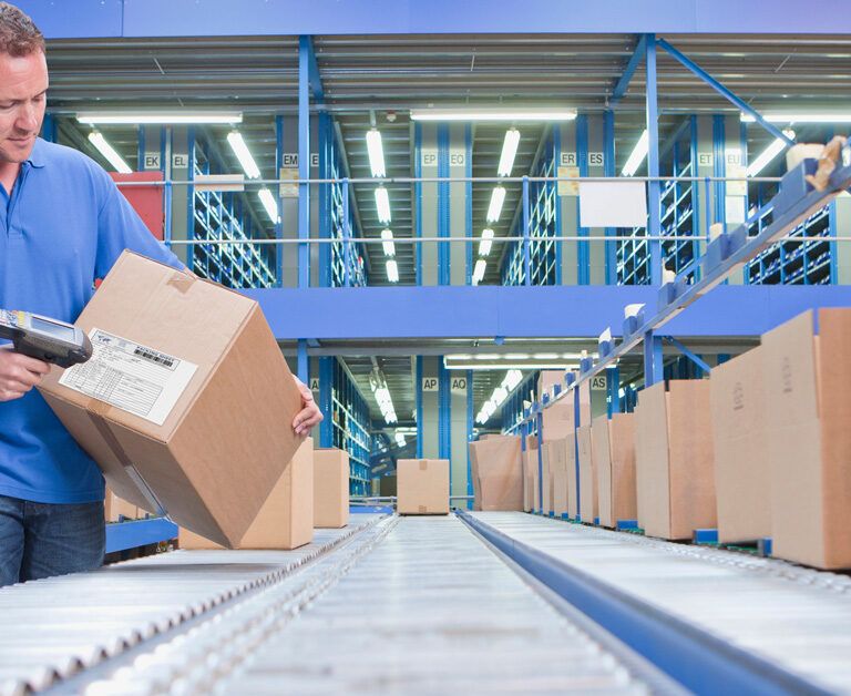 A man in a blue shirt scans a shipping label on a cardboard box in a warehouse, standing beside a conveyor belt with several other boxes—a scene streamlined by distributed order management under bright industrial lights.
