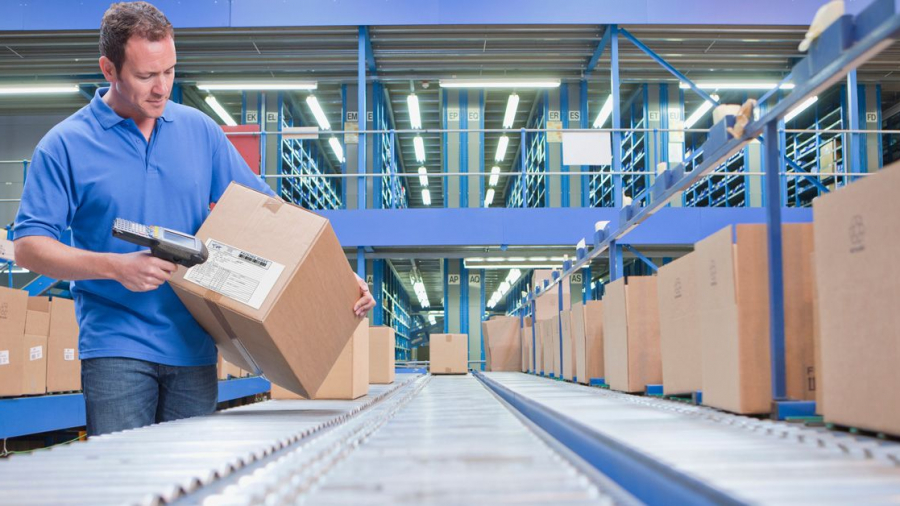 A man in a blue shirt scans a shipping label on a cardboard box in a warehouse, standing beside a conveyor belt with several other boxes—a scene streamlined by distributed order management under bright industrial lights.