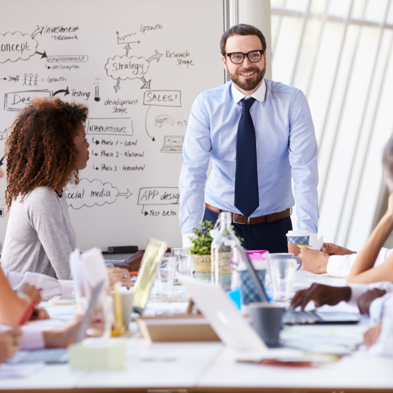 A man in a shirt and tie stands smiling in front of a whiteboard covered in notes, leading a meeting with five colleagues seated around a table in a brightly lit office.
