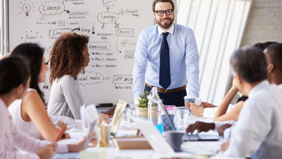 A man in a shirt and tie stands smiling in front of a whiteboard covered in notes, leading a meeting with five colleagues seated around a table in a brightly lit office.
