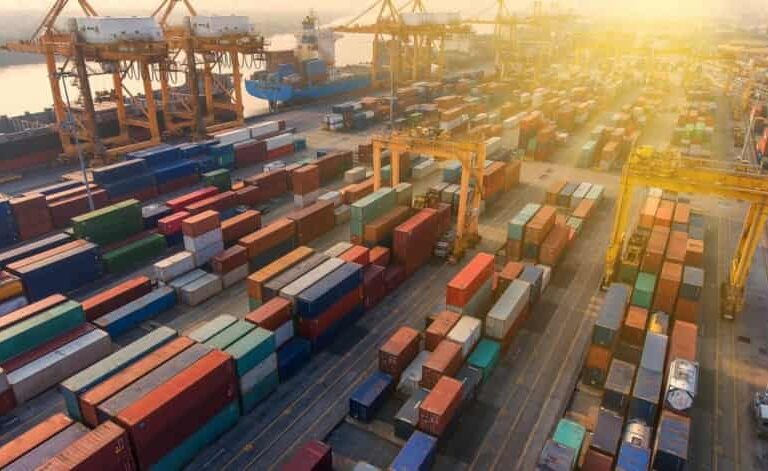 Aerial view of a busy shipping port with numerous colorful stacked cargo containers, large cranes, and ships docked by the water under bright sunlight.