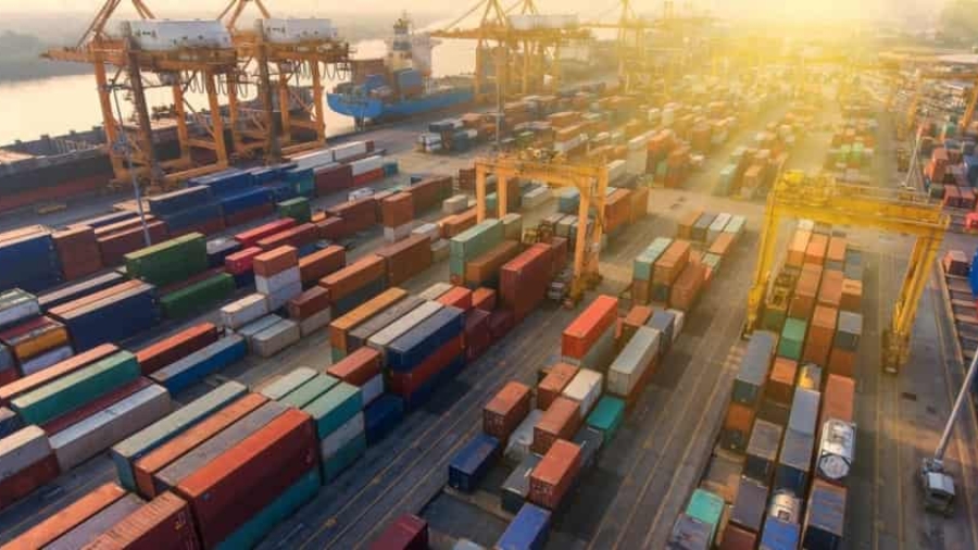 Aerial view of a busy shipping port with numerous colorful stacked cargo containers, large cranes, and ships docked by the water under bright sunlight.