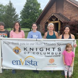 Six people stand outside a brick building, holding a banner thanking Knights of Columbus, enVista, and Aldea Wellness for their community impact as sponsors of a Haiti ministry. Grass and trees can be seen in the background.