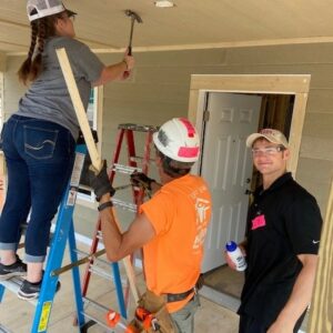 Three people work on a house porch, showcasing community impact; a woman uses a hammer on a ladder, assisted by a man in an orange shirt and hard hat, while another stands nearby holding a water bottle and smiling at the camera.