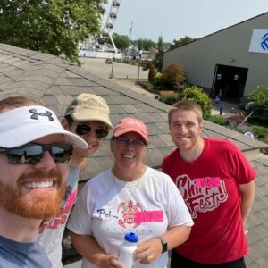Four people smiling and posing for a selfie on a sunny rooftop, with a Ferris wheel, trees, and buildings in the background. Their cheerful gathering highlights community impact in a relaxed, casual setting.