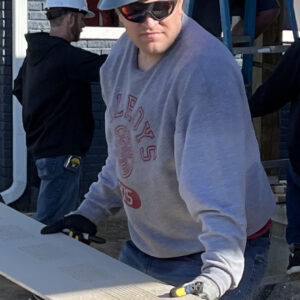 A person wearing a hard hat, sunglasses, gloves, and a gray sweatshirt carries a long wooden board at a construction site, contributing to the community impact. Other workers and a ladder can be seen in the background.