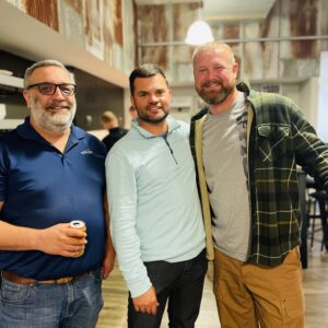 Three men stand indoors, smiling together and enjoying a drink. Casually dressed against rustic, corrugated metal walls and wooden floors, they embody a relaxed, friendly atmosphere that reflects strong community impact.