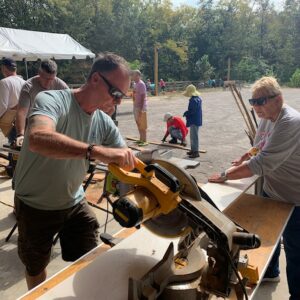 A man uses a circular saw to cut wood on a workbench outdoors, contributing to the community impact as others focus on carpentry projects around him. Everyone works together with tools and materials under a sunny sky.
