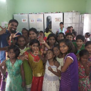 A group of smiling children and a man pose together in a brightly lit room with lockers and green walls, showcasing community impact. Most of the children wear colorful clothes and make peace signs or playful gestures.