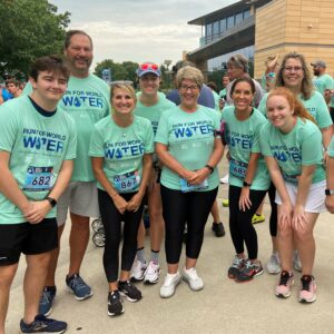 A group of smiling people in matching Run For World Water shirts stand together outdoors before a race, showcasing their community impact as they wear running shoes and bib numbers, with other participants and a building in the background.