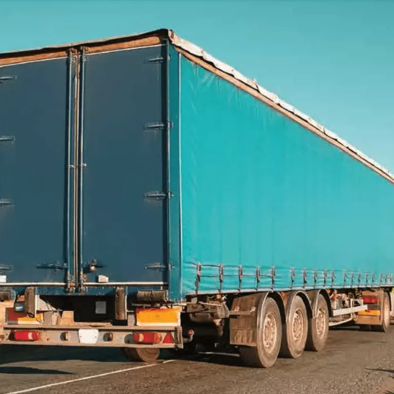 A large blue freight truck with a covered trailer is driving down an empty road under a clear blue sky, with dry grassland visible in the background.