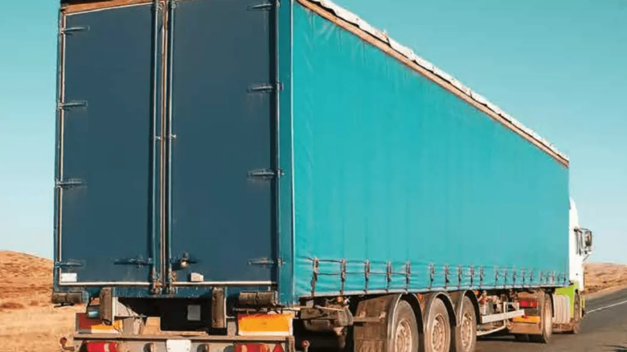A large blue freight truck with a covered trailer is driving down an empty road under a clear blue sky, with dry grassland visible in the background.
