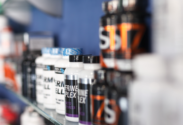 Several bottles of dietary supplements are lined up on a glass shelf, with labels facing forward. The background is blurred, highlighting the bottles in the foreground against a blue wall.