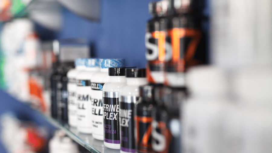 Several bottles of dietary supplements are lined up on a glass shelf, with labels facing forward. The background is blurred, highlighting the bottles in the foreground against a blue wall.