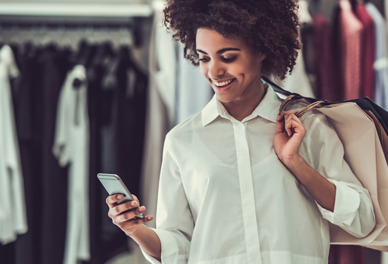 A woman holding shopping bags smiles while looking at her smartphone in a clothing store, with racks of clothes visible in the background.