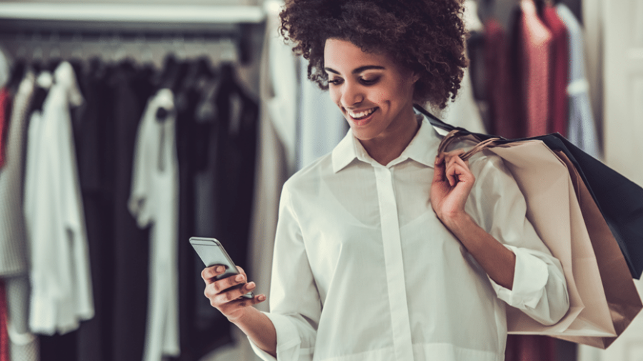 A woman holding shopping bags smiles while looking at her smartphone in a clothing store, with racks of clothes visible in the background.