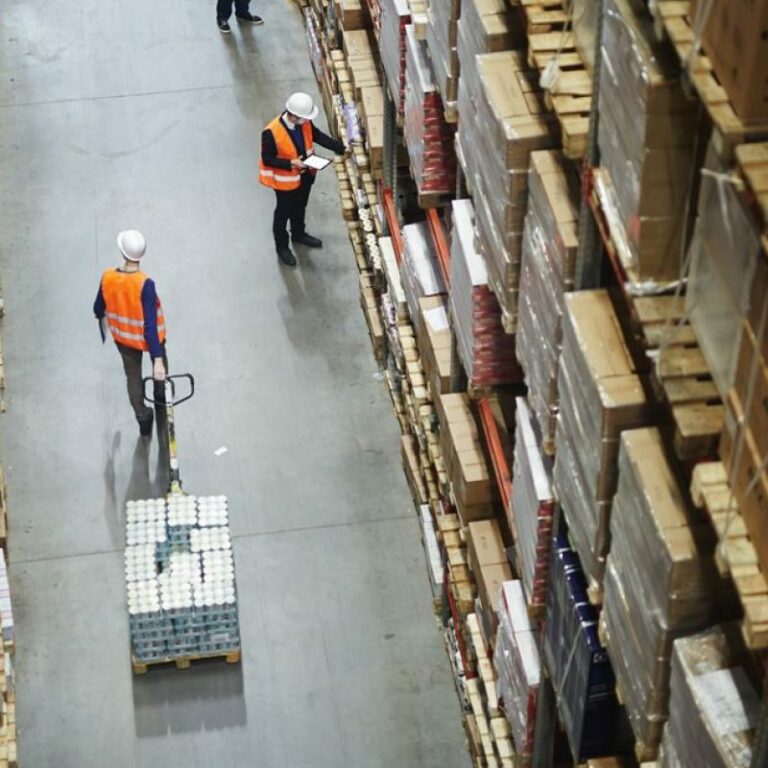 Aerial view of a warehouse aisle with two workers in safety vests and helmets; one is moving a pallet with a hand truck, while the other checks a clipboard among tall shelves stacked with boxes.