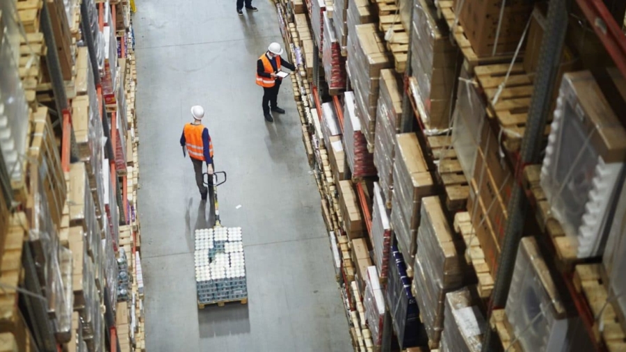Aerial view of a warehouse aisle with two workers in safety vests and helmets; one is moving a pallet with a hand truck, while the other checks a clipboard among tall shelves stacked with boxes.
