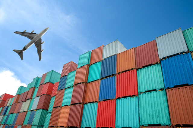 A commercial airplane flies above a large stack of colorful shipping containers under a clear blue sky.