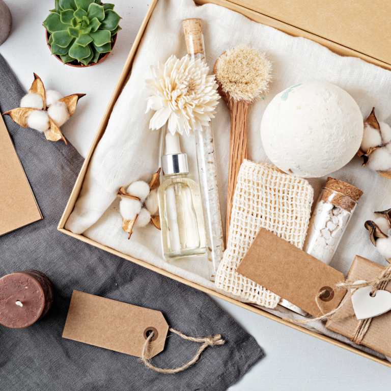 A gift box with bath and body items including a bath bomb, soap, dried flowers, glass bottles, and a scrubber, arranged neatly with brown tags and cotton flowers. Nearby are candles, a plant, and blank cards.