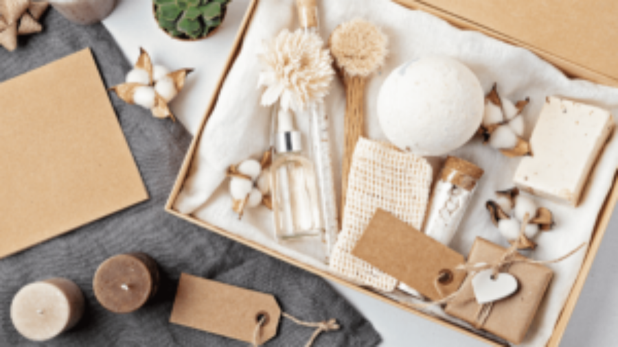 A gift box with bath and body items including a bath bomb, soap, dried flowers, glass bottles, and a scrubber, arranged neatly with brown tags and cotton flowers. Nearby are candles, a plant, and blank cards.